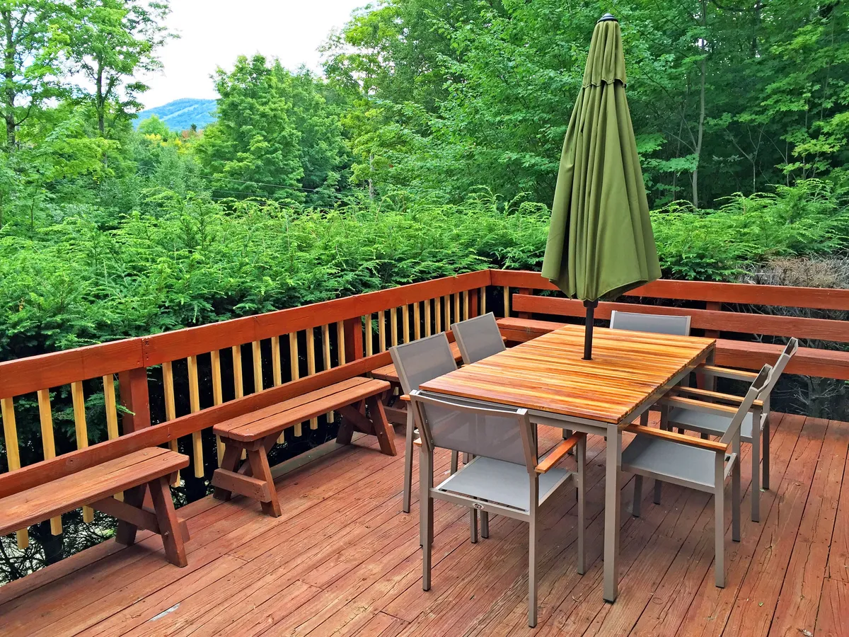 The image shows an outdoor dining area with a green umbrella, wooden benches, and a table surrounded by lush greenery. The setup invites families to gather for meals or games outside, enjoying fresh mountain air and nature views. The communal arrangement and scenic backdrop make it ideal for group enjoyment.