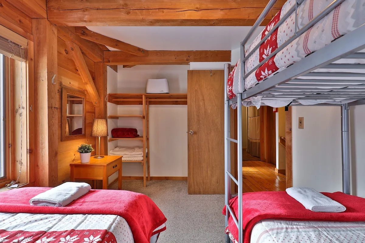 A cozy bedroom featuring a bunk bed with sturdy ladders and an adjacent single bed, accented by wooden beams, a lamp, and a practical shelf. The space is bathed in natural light, with inviting linens and warm wood tones creating a welcoming family environment.