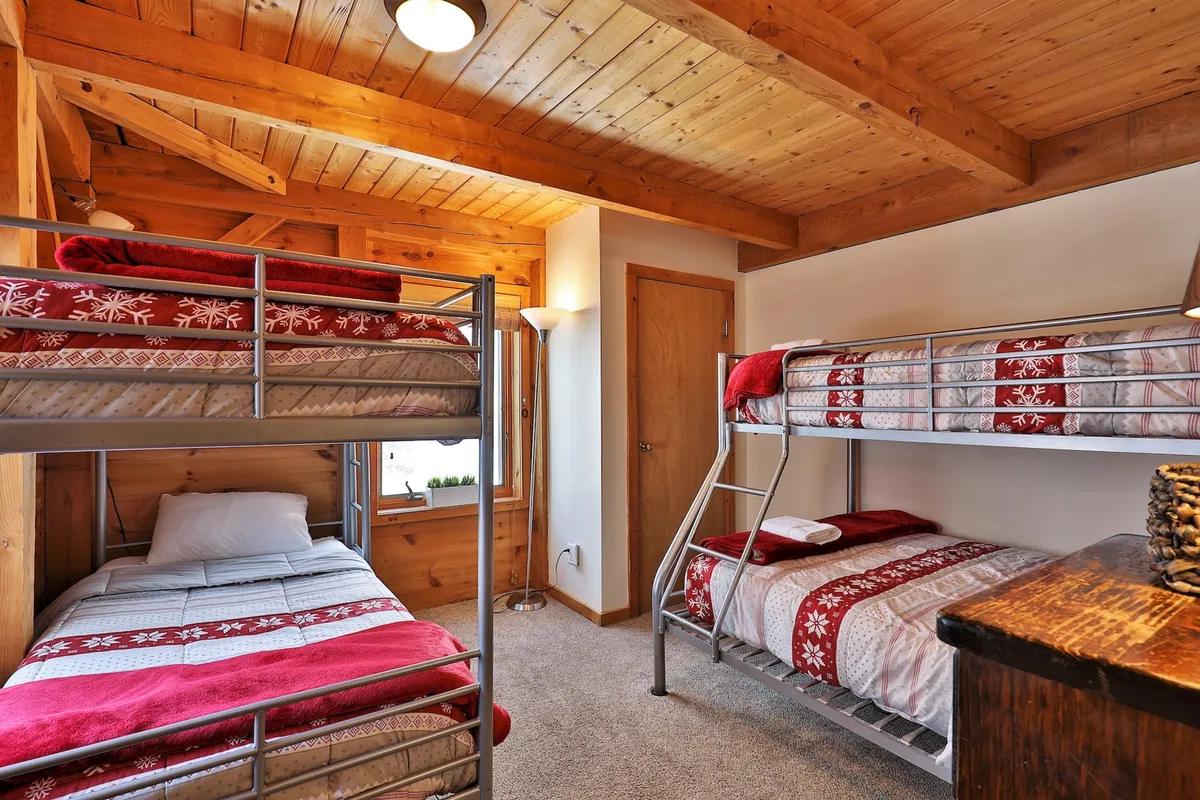 This image features a bright and inviting bunk bedroom, with wooden ceilings, decorative bedding, a lamp, and wooden furniture. The bunk beds are clearly visible, drawing attention to their design and suitability for families and groups.