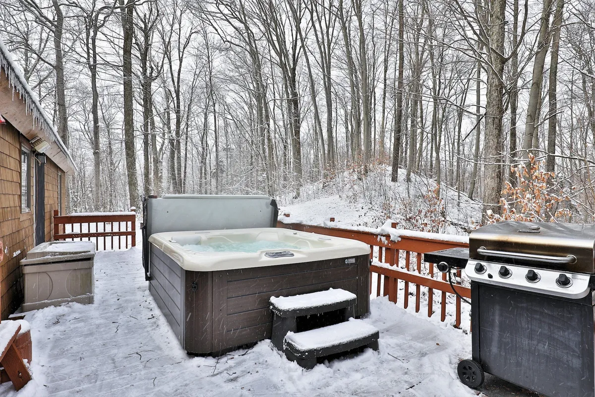 A snow-covered deck featuring a hot tub and grill, bordered by wooden railings and surrounded by winter trees. The scene captures an inviting space for relaxing soaks, outdoor grilling, and gathering with friends or family, all while enjoying the crisp Vermont air.