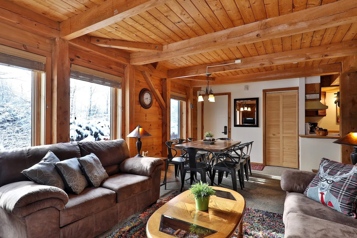 This image showcases the heart of the cabin's living space: a warm and inviting open-plan area with exposed wooden beams, large windows that bathe the space in natural light, a central dining table, and plush sofas. Decorative pillows and rustic accents add to the cozy, communal feel, and the layout encourages gathering for family meals, game nights, or conversation.