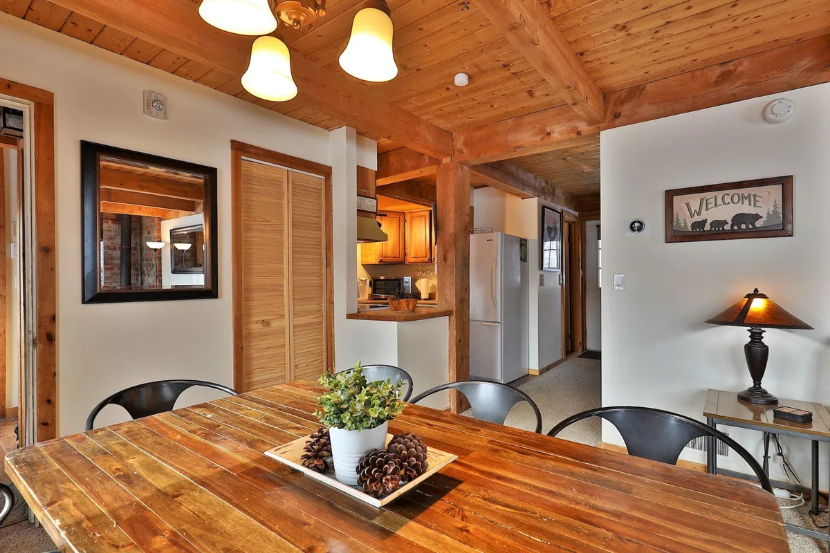 The image features a wooden dining table surrounded by chairs and a welcoming sign, with a clear view into the adjacent kitchen area. The layout highlights the seamless flow between dining and kitchen, reinforcing the open-concept design. Soft lighting and wood elements make the space feel both homey and functional for shared meals or casual gatherings.