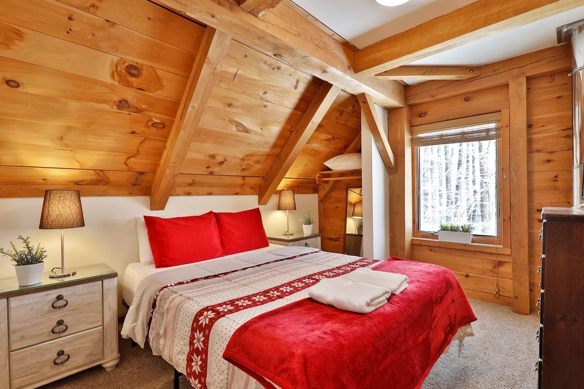 A tranquil adult bedroom with a queen bed dressed in red and white linens, warm wooden beams overhead, a window offering views of the surrounding trees, and a nightstand with a reading lamp.