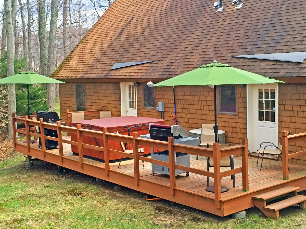 A spacious deck featuring a hot tub, grill station, green umbrellas, and wooden railing, surrounded by greenery. The deck is set up for outdoor dining, relaxation, and social gatherings, with clear invitations for grilling and stargazing after a day’s adventure.