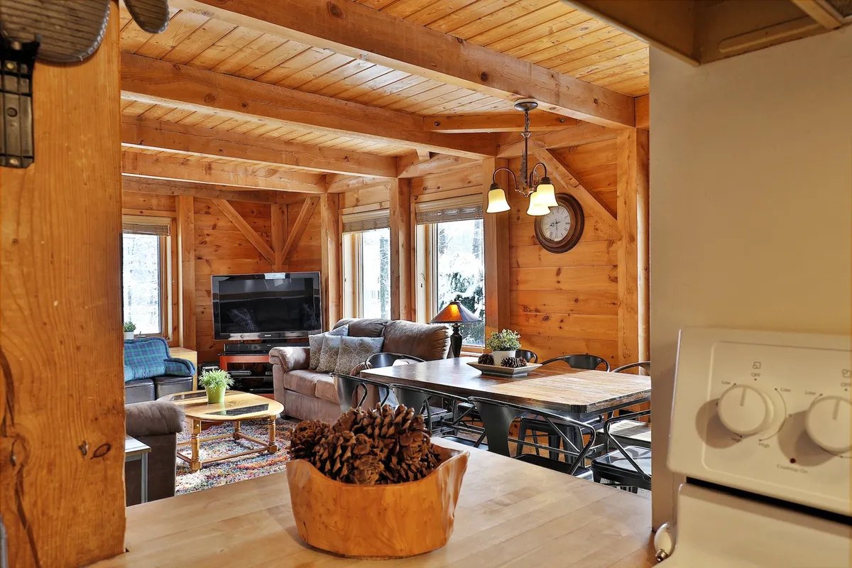 An inviting open-plan living area with exposed wooden beams, a comfortable sofa facing a fireplace, a large dining table, and expansive windows that flood the space with natural light and scenic views. The space is arranged to facilitate group gatherings, game nights, and cozy socializing.