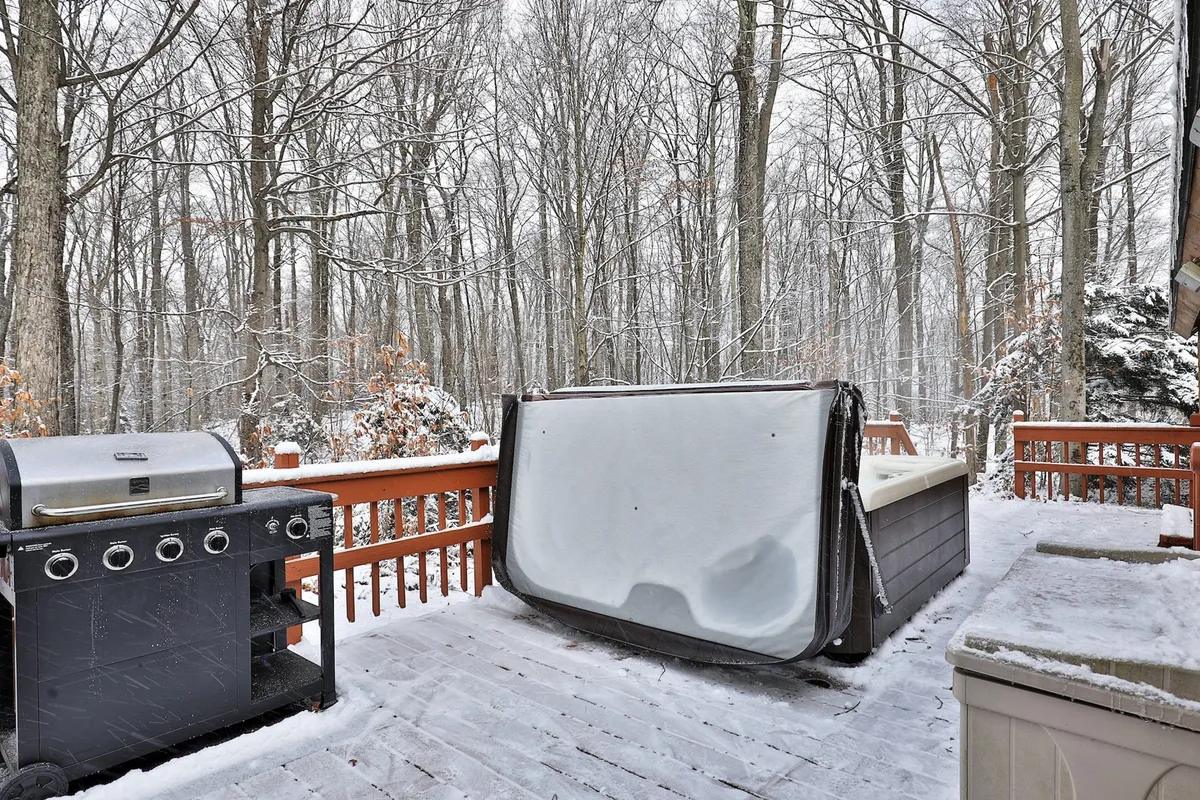 A peaceful winter deck scene with a grill, hot tub, and wooden railing, framed by snow-laden trees. The warm steam from the hot tub contrasts with the cool outdoor environment, and the setting sun hints at tranquil evenings spent outside.