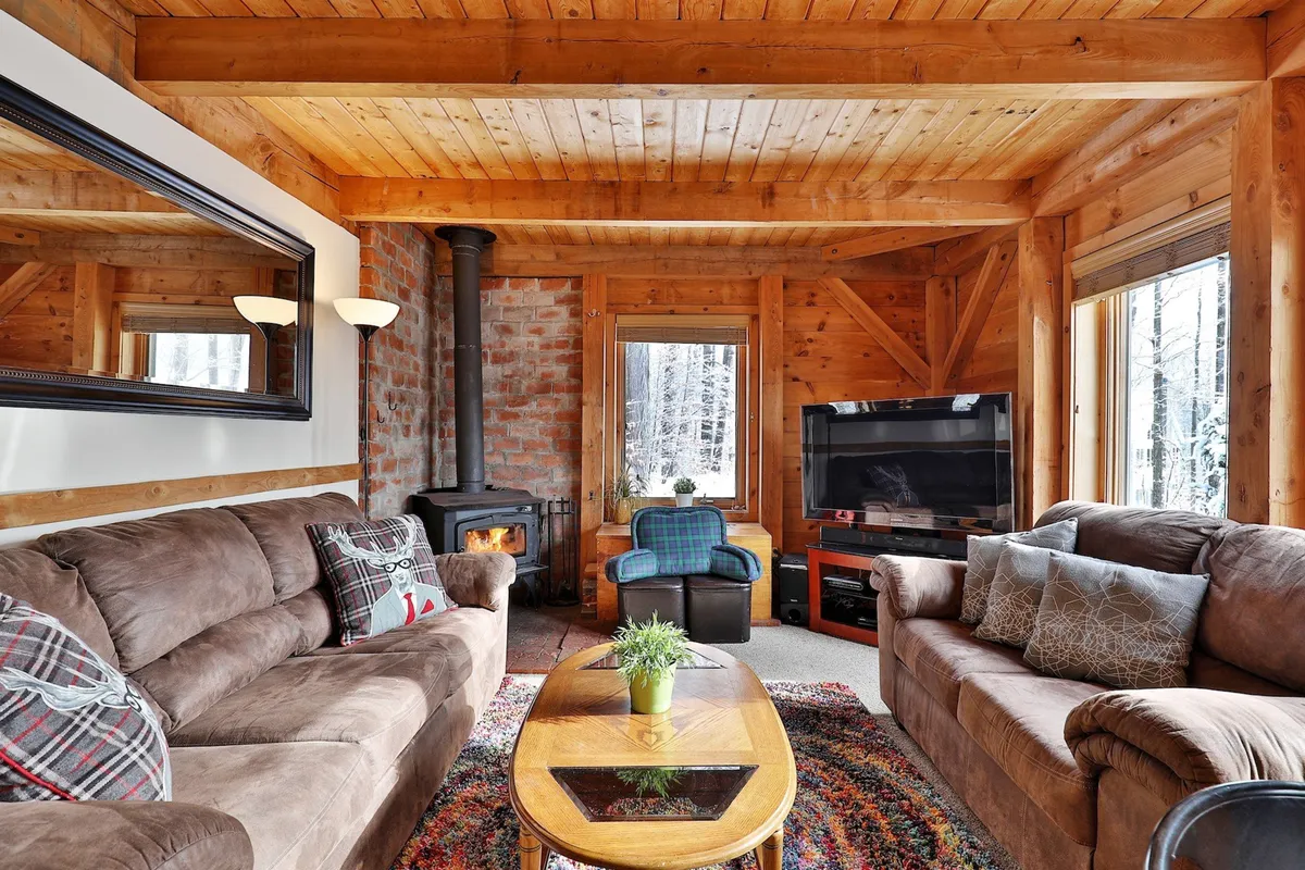 The hero image showcases a cozy living area with exposed wooden beams, a crackling fireplace, plush sofas, and large windows revealing a snowy Vermont landscape. The setting feels warm and inviting, perfect for gathering with family or friends after a day of mountain adventures.