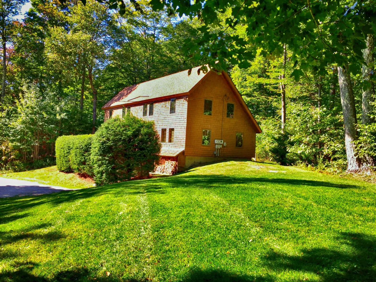 The image displays the wooden exterior of the cabin, set amidst manicured lawns and tall trees. The natural landscape, along with the inviting porch, communicates proximity to nature and an authentic mountain retreat atmosphere, ideal as a ski basecamp.