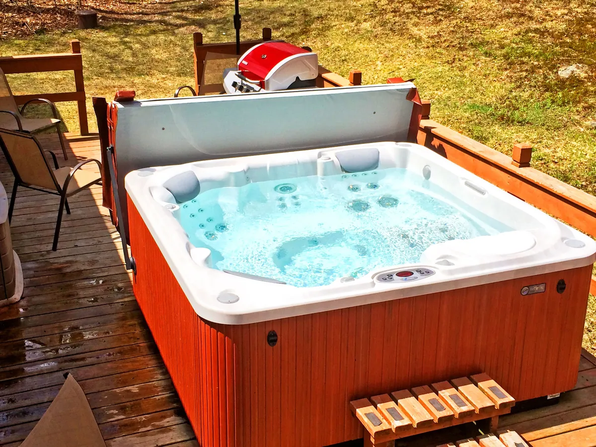 This image showcases the exterior hot tub set on a deck, with a grill also visible. The tranquil outdoor setting is designed for relaxation and socializing, and the hot tub is a focal point for group gatherings and celebration, set against the backdrop of Vermont’s natural beauty.