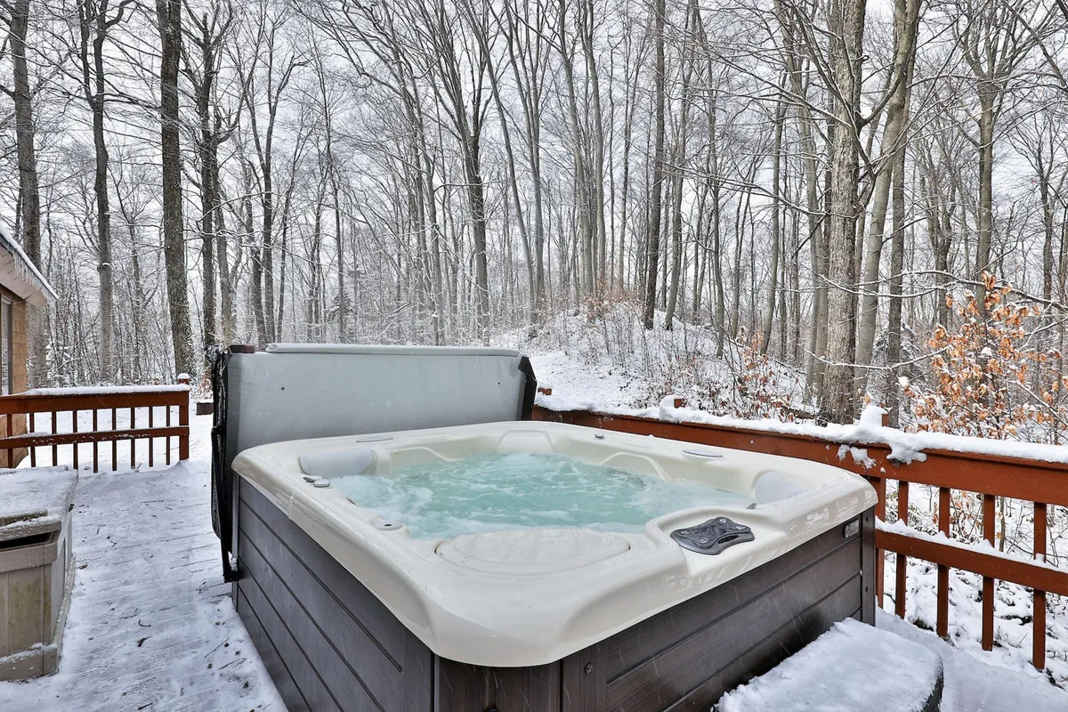 This exterior image features a private hot tub set on a deck surrounded by trees, with a picturesque winter backdrop. The scene is tranquil and inviting, perfect for relaxing after a day on the slopes. Steam rises from the tub, hinting at warmth and comfort despite the winter chill.